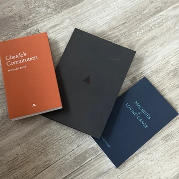 A tabletop photograph of three clothbound technology books: an orange “Claude’s Constitution, January 2026” volume marked “AI,” a black Vercel employee manual distributed only to Vercel employees, and a blue “Machines of Loving Grace” volume credited to Dario Amodei. The source post calls them “The 3 most important books of modern technology.”