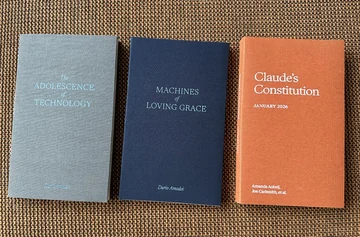 A photograph of three clothbound AI essay volumes arranged on a brown sofa: gray “The Adolescence of Technology” by Dario Amodei; navy “Machines of Loving Grace” by Dario Amodei; and orange “Claude’s Constitution, January 2026” by Amanda Askell, Joe Carlsmith, et al. The source caption reads “Gotta collect em all.”