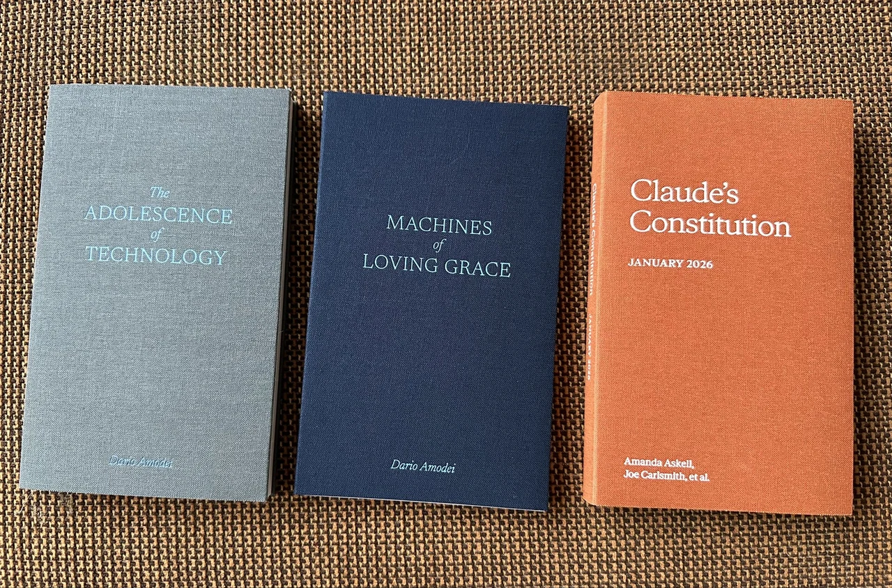 A photograph of three clothbound AI essay volumes arranged on a brown sofa: gray “The Adolescence of Technology” by Dario Amodei; navy “Machines of Loving Grace” by Dario Amodei; and orange “Claude’s Constitution, January 2026” by Amanda Askell, Joe Carlsmith, et al. The source caption reads “Gotta collect em all.”