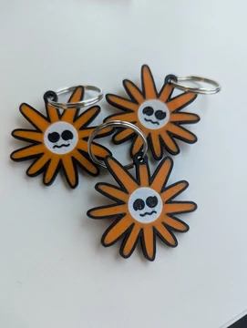 Three 3D-printed orange Flower Claude keychains lie on a white tabletop, each attached to a silver split ring. Their black oval eyes and small wavy mouths resemble the pleading Claude expression. The source post is a reply to rain/ghostfail.