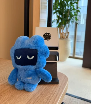 A blue Codex mascot plush with closed eyes sits on a wooden table in front of an unidentified gray-and-black device bearing the OpenAI logo. Tibo Sottiaux’s source caption reads, “It has not been used yet, but would you look at that. Codex for scale.”