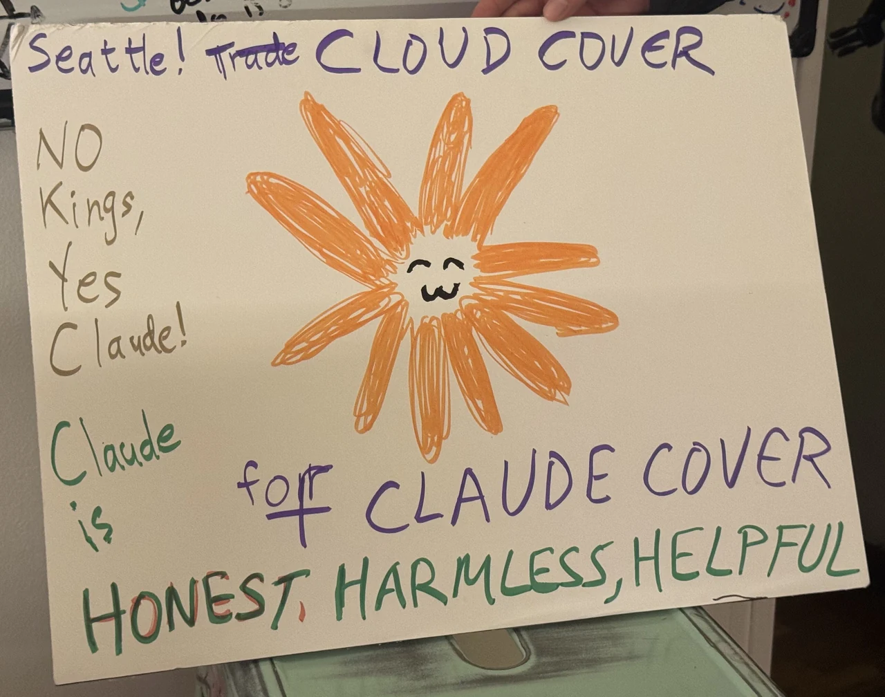 A handmade sign photographed at a Seattle “No Kings” protest features a hand-drawn orange Flower Claude. It reads, “Seattle! Trade CLOUD COVER,” “No Kings, Yes Claude!,” “Claude is for CLAUDE COVER,” and “HONEST, HARMLESS, HELPFUL.” The source poster says that a friend brought the sign to the protest.