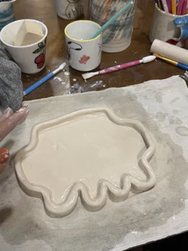 Ann Nguyen’s Clawd-shaped pottery tray in progress on a workshop table. The pale clay piece has a shallow center, a raised rim, rounded side protrusions, and short feet along its lower edge. It rests on a cloth beside decorated mugs, brushes, pottery tools, and a sponge; a clay-covered hand is visible at the left. There is no readable text on the tray. Ann’s source post says, “missed the plushies so i decided to make my own pottery claudie” and “hope it turns out well”, followed by a laughing emoji. The post quotes a Claude plushie giveaway and also includes an image of an orange version of the tray.