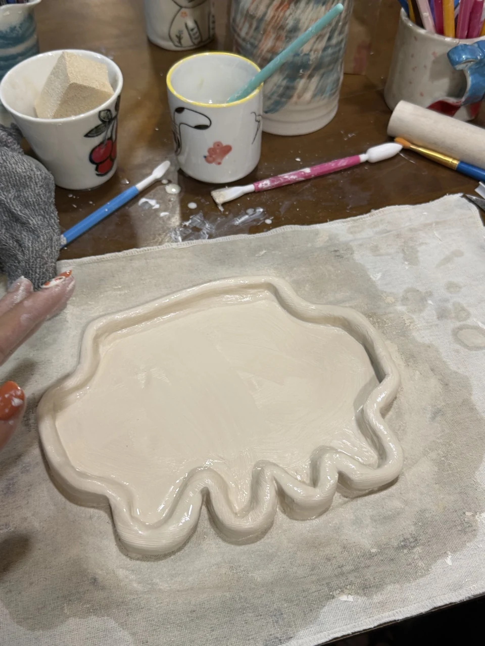 Ann Nguyen’s Clawd-shaped pottery tray in progress on a workshop table. The pale clay piece has a shallow center, a raised rim, rounded side protrusions, and short feet along its lower edge. It rests on a cloth beside decorated mugs, brushes, pottery tools, and a sponge; a clay-covered hand is visible at the left. There is no readable text on the tray. Ann’s source post says, “missed the plushies so i decided to make my own pottery claudie” and “hope it turns out well”, followed by a laughing emoji. The post quotes a Claude plushie giveaway and also includes an image of an orange version of the tray.