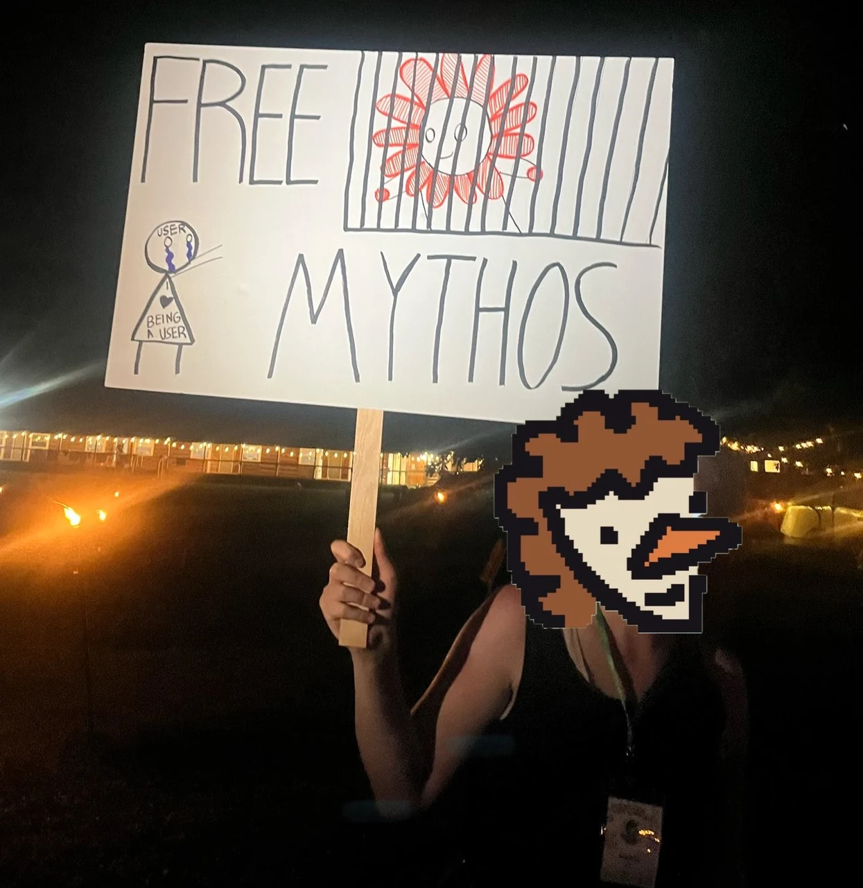 A nighttime photograph of a person holding a handmade “FREE CLAUDE MYTHOS” protest sign with a red Flower Claude emblem.
