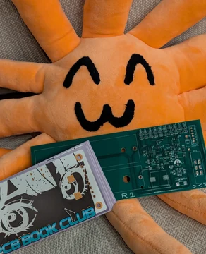 An orange Claude plush rests beside a printed circuit board and a small screen. The source calls the plush Haiku and says, “haiku loves pcbs.”