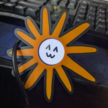 A physical orange-and-gray Flower Claude ornament or badge stands in front of a computer keyboard. Source caption: “i felt compelled to give Claude a physical form.”