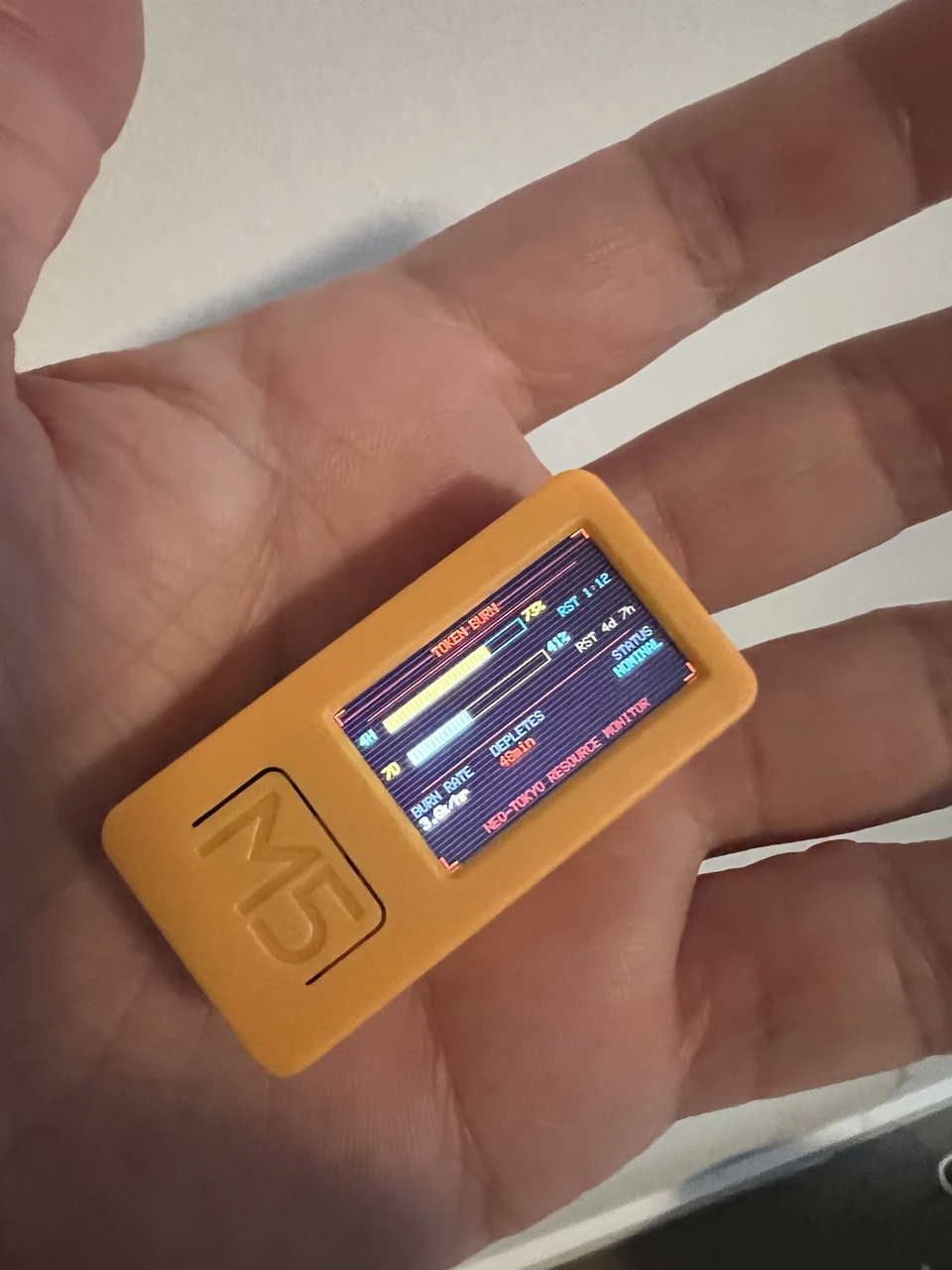 A hand holds an orange M5StickC Plus2 running a custom Claude token-usage burn monitor over Wi-Fi.