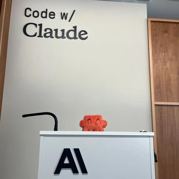 An official Claude plush sits at a podium beneath the sign “Code w/ Claude” at Code with Claude San Francisco.