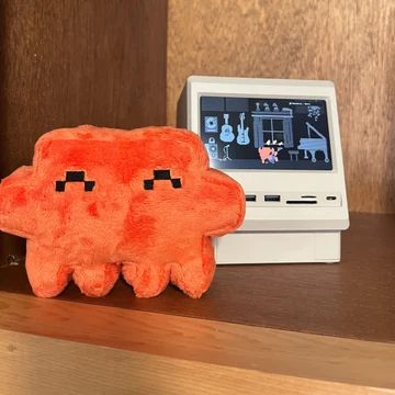 An official Claude plush poses beside a miniature computer displaying pixel-art musicians at Code with Claude San Francisco.