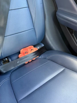 An official Claude plush is tucked into a blue car seat with the seat belt fastened across it.