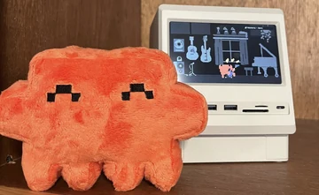 An official Claude plush stands beside a miniature computer whose screen shows a pixel-art music room.
