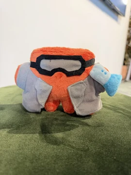 An official Claude plush wears dark goggles and a gray vest while holding a blue laboratory flask at a Code with Claude demo booth.