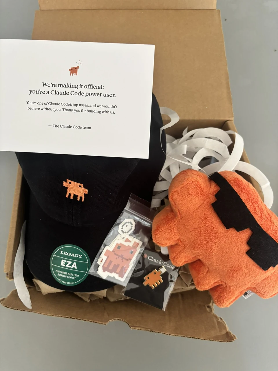 An open Claude Code gift box contains an orange plush, black cap, pixel stickers, pin, and a card reading: “We’re making it official: you’re a Claude Code power user.”
