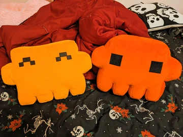Two orange Claude plushes, one yellow-orange and one red-orange, lie together on patterned bedding.