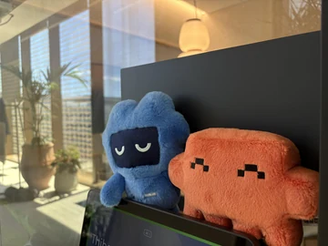 A blue Codex mascot plush with closed eyes and an orange Clawd plush with pixel eyes sit together atop a monitor in a sunlit OpenAI office. Tibo Sottiaux’s source caption reads, “Spotted Codex making friends at the OpenAI office.”