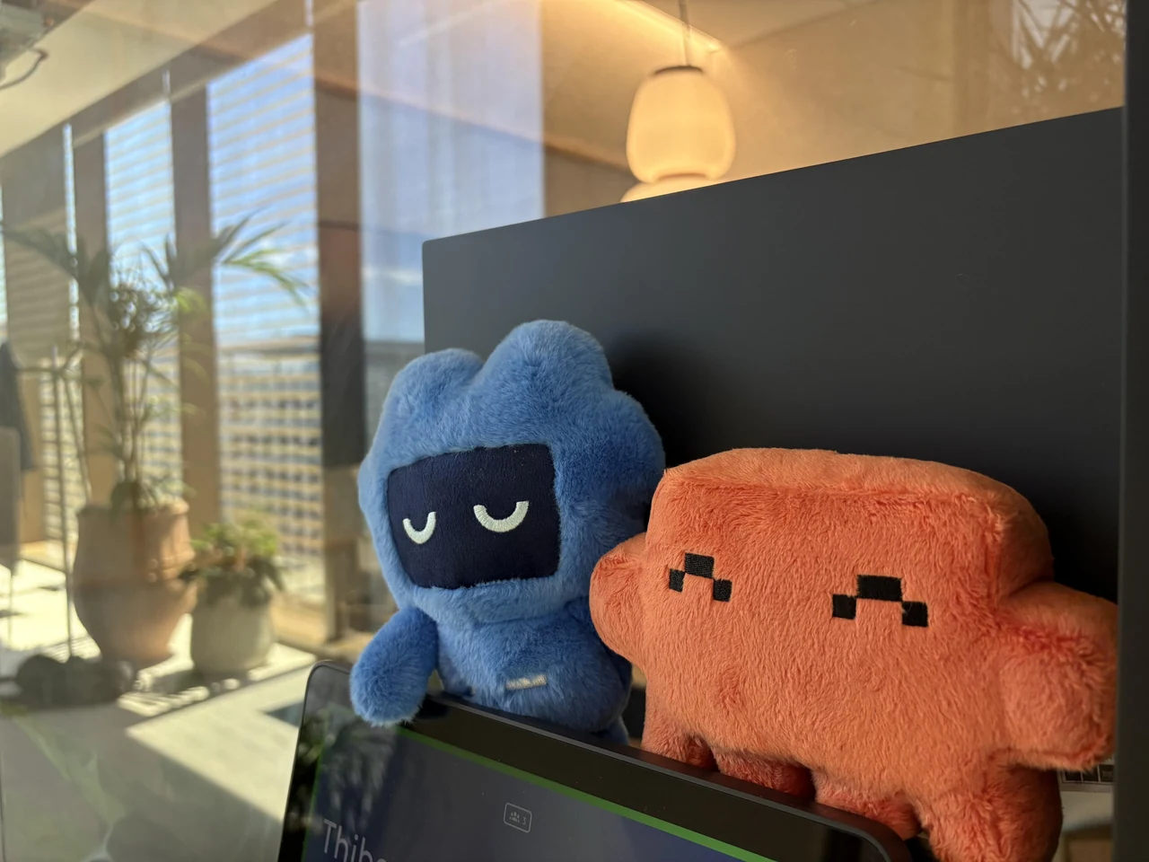 A blue Codex mascot plush with closed eyes and an orange Clawd plush with pixel eyes sit together atop a monitor in a sunlit OpenAI office. Tibo Sottiaux’s source caption reads, “Spotted Codex making friends at the OpenAI office.”